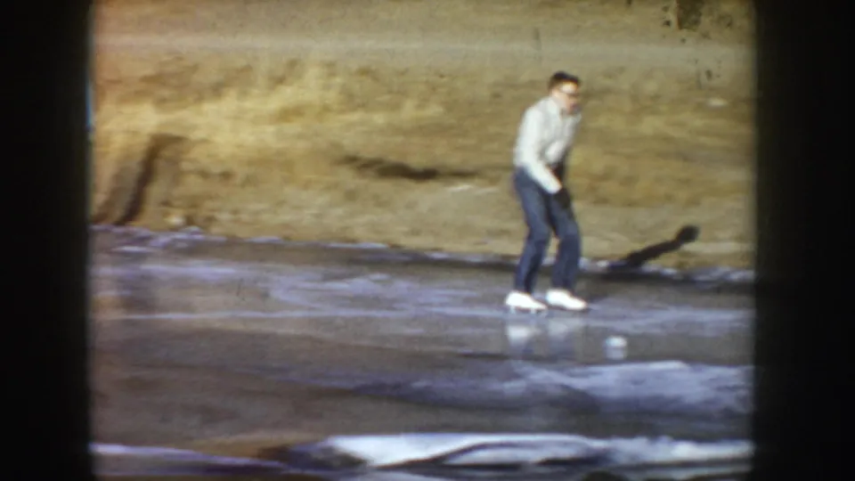 Young man skating on a natural frozen pond in what may be Idaho, circa 1955; white sweater, dark jeans, white ice skates; 8mm home movie frame.