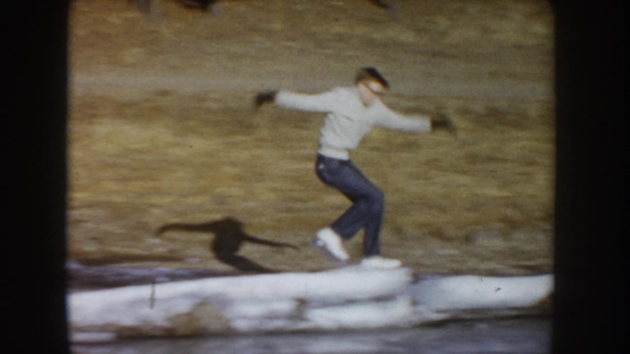 Young man spreading arms for balance while skating on a frozen outdoor pond, circa 1955; white sweater, dark jeans; 8mm color home movie frame.