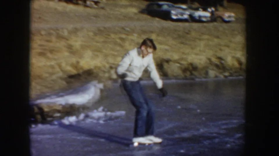 A skater leaning forward on frozen ice with a dark-toned mid-century automobile parked on the bank behind; Idaho, circa 1955; 8mm home movie frame.