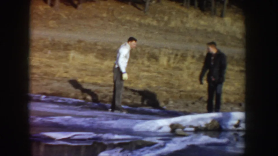 Two men, one in a white shirt and one in darker clothing, standing at the margin of a frozen outdoor pond; Idaho, circa 1955; 8mm home movie frame.