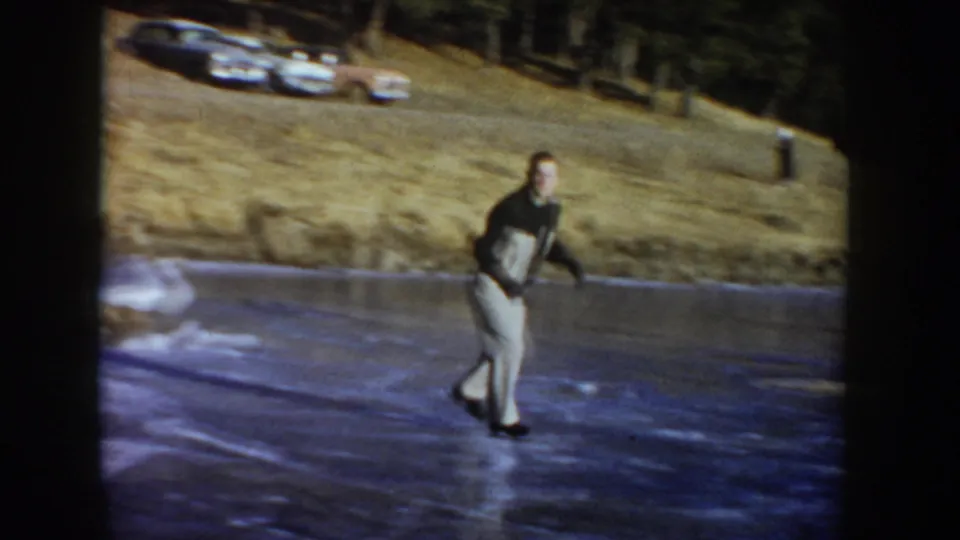 In 1955 Idaho home-movie footage, a young man cautiously skates on a frozen pond below a grassy bank with mid-century cars.