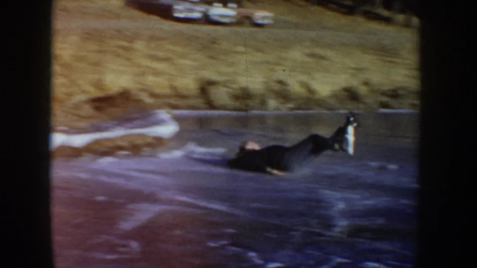 The young skater lies on the frozen pond after a fall, one leg lifted against the soft, grainy 8mm image.