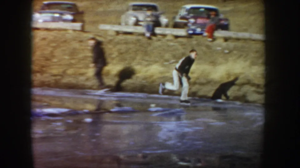 The skater moves across the frozen pond in Idaho, with a dog, a standing spectator, and 1950s cars visible on the hillside.