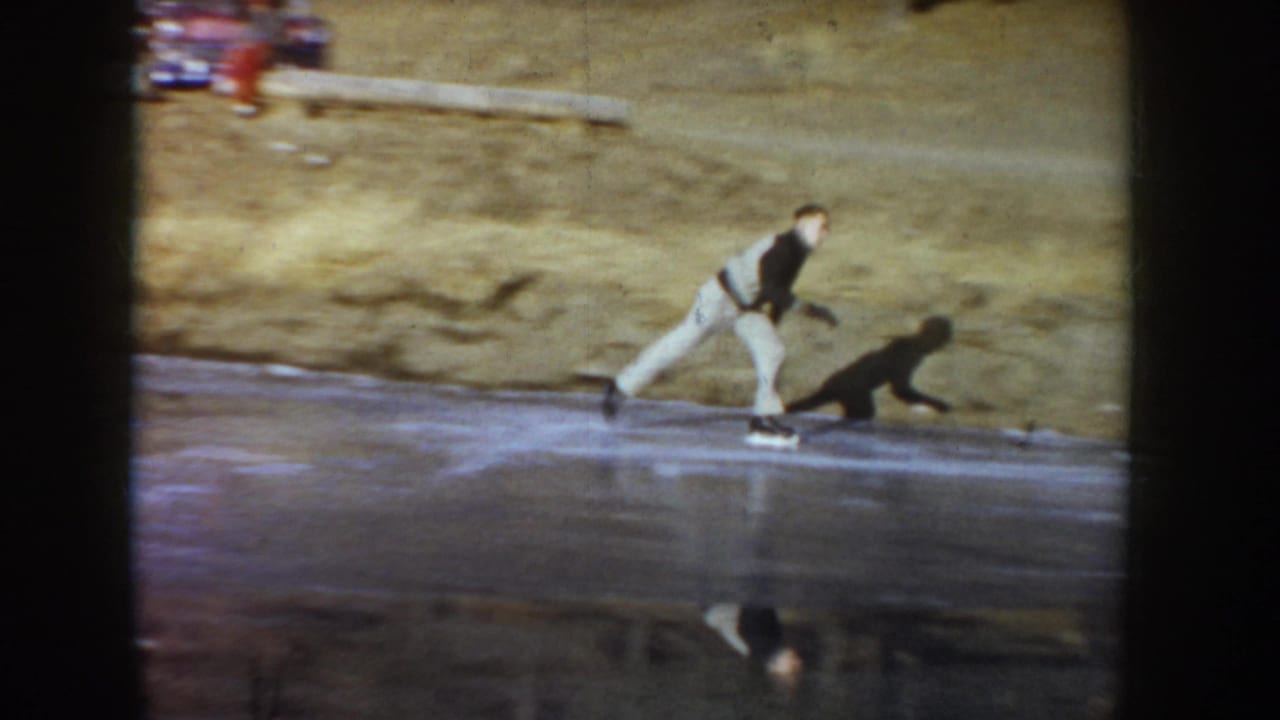Man and dog on a frozen rural pond, Idaho, circa 1955; skater leans into stride as dog runs at his side on reflective ice with a grassy winter bank behind.