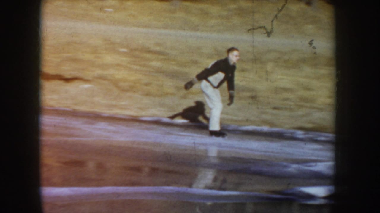 Man in dark jacket skating upright on frozen pond, dark dog visible to his lower right, grassy slope behind, warm color shift from aged 8mm film.