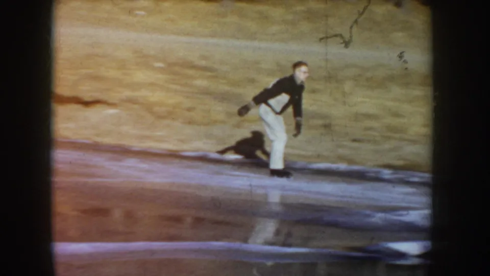Man skating upright on a frozen rural pond, Idaho, circa 1955, dark dog running alongside; warm reddish color cast and grain from aged 8mm reversal stock.