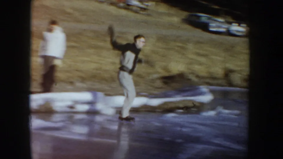 Skater on frozen pond, Idaho, circa 1955, arms raised mid-glide; a second person in white stands at the left edge and a dark parked vehicle is visible on the bank.