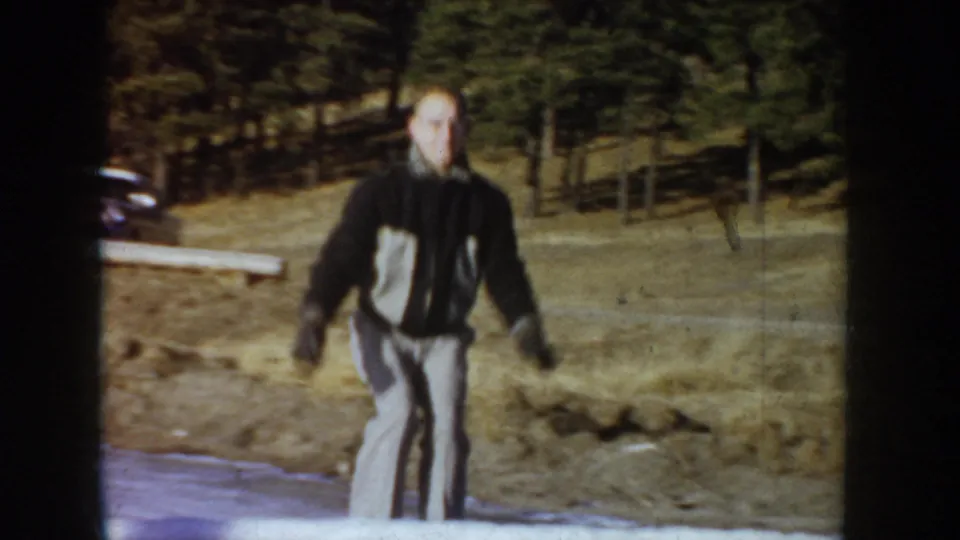 Close view of man on ice skates facing the camera, Idaho, circa 1955; wooden fence post and conifer-covered slope behind, face clearly visible.