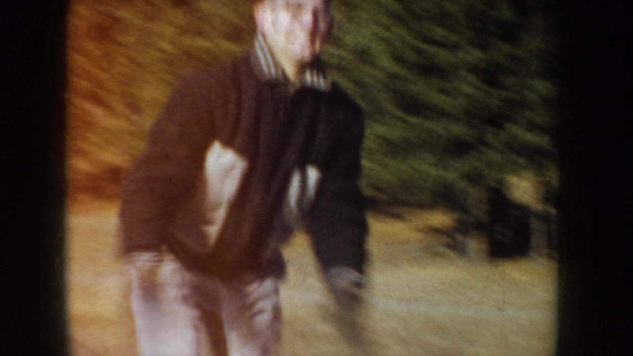 Man in dark knit sweater and scarf gliding on an outdoor frozen pond, Idaho, circa 1955; conifer trees in background; 8mm home movie footage.