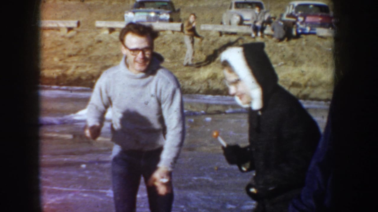 Candid 8mm frame, circa 1955 Idaho: man in horn-rimmed glasses and gray sweater and a woman in a dark coat and white head covering laughing together outdoors near parked mid-century automobiles.
