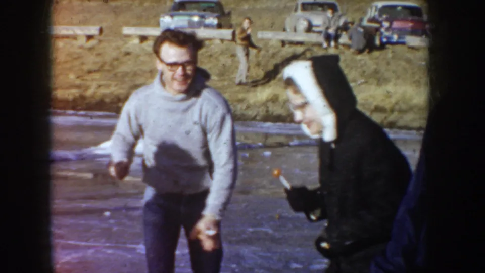 Candid 8mm frame, circa 1955 Idaho: man in horn-rimmed glasses and gray sweater and a woman in a dark coat and white head covering laughing together outdoors near parked mid-century automobiles.