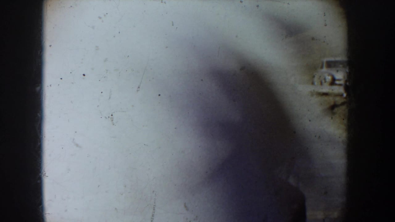 Film damage frame from a circa 1955 8mm home movie reel: extreme overexposure with white bleach-out, partial dark shadow, faint vehicle outline, and two blue U-shaped marks drawn directly on the film stock.