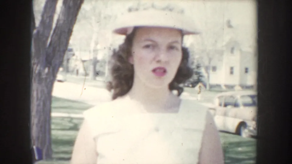 Close-up portrait of a young woman in a white summer hat and sleeveless white top, with a light-colored vintage car and residential building in the background, circa 1955.
