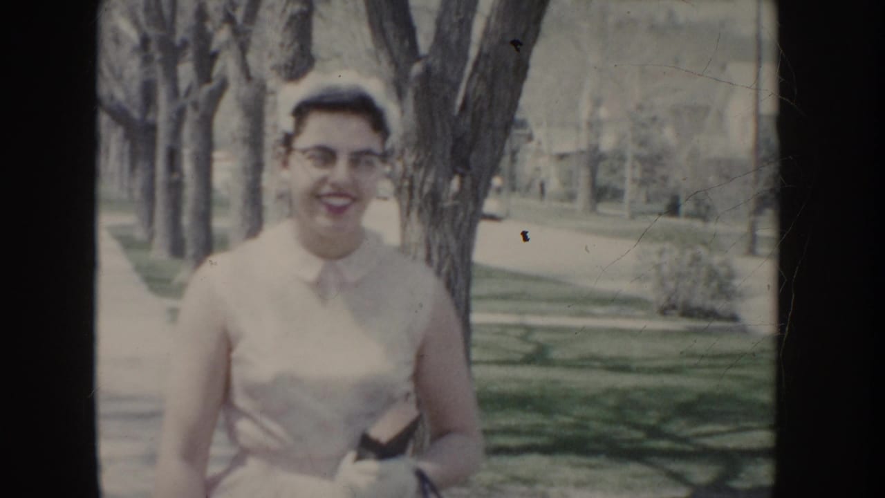Young woman wearing white sleeveless dress, cat-eye glasses, and small white cap smiling at camera in a tree-lined suburban setting, Idaho, c. 1955.