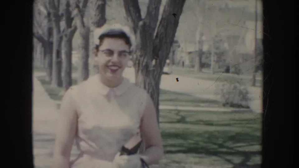 Young woman wearing white sleeveless dress, cat-eye glasses, and small white cap smiling at camera in a tree-lined suburban setting, Idaho, c. 1955.
