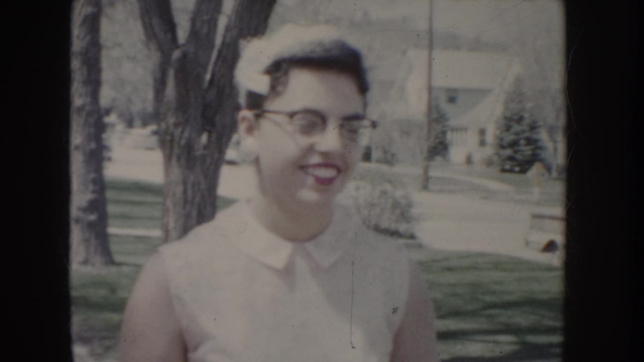 Young woman in white dress, cat-eye glasses, and white cap smiling in medium shot, suburban house and trees visible behind her, c. 1955.