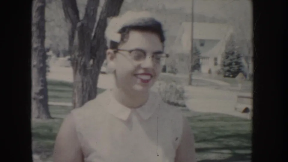 Young woman in white dress, cat-eye glasses, and white cap smiling in medium shot, suburban house and trees visible behind her, c. 1955.