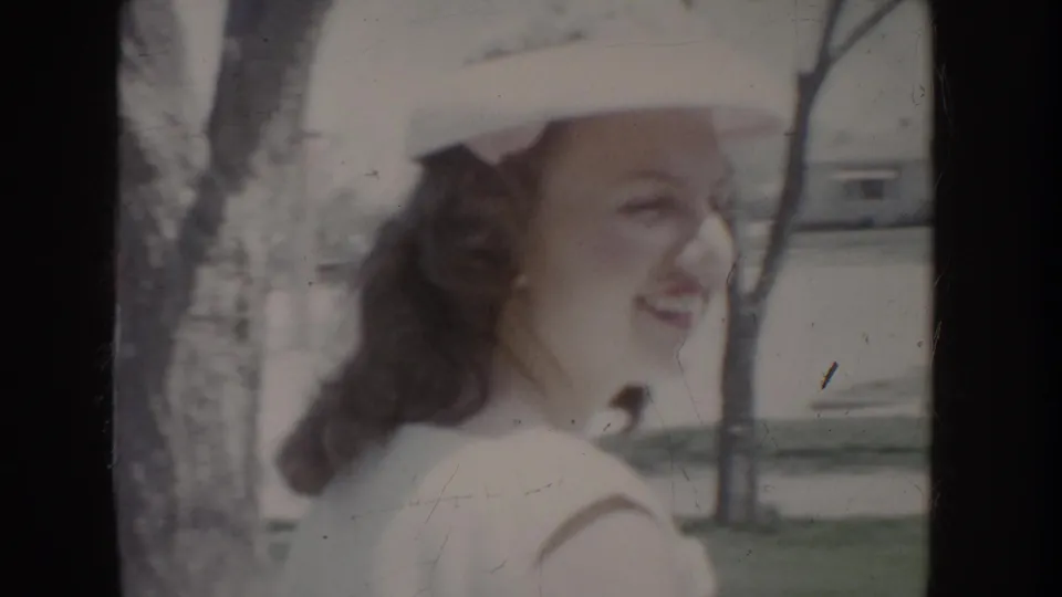 Young woman in wide-brimmed white hat and white dress shown in three-quarter profile, smiling, with trees in suburban background, c. 1955.
