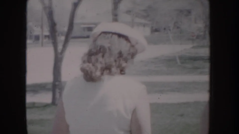 Young woman in white dress and wide-brimmed white hat viewed from behind in three-quarter angle, suburban residential setting visible behind her, 8mm home movie c. 1955.