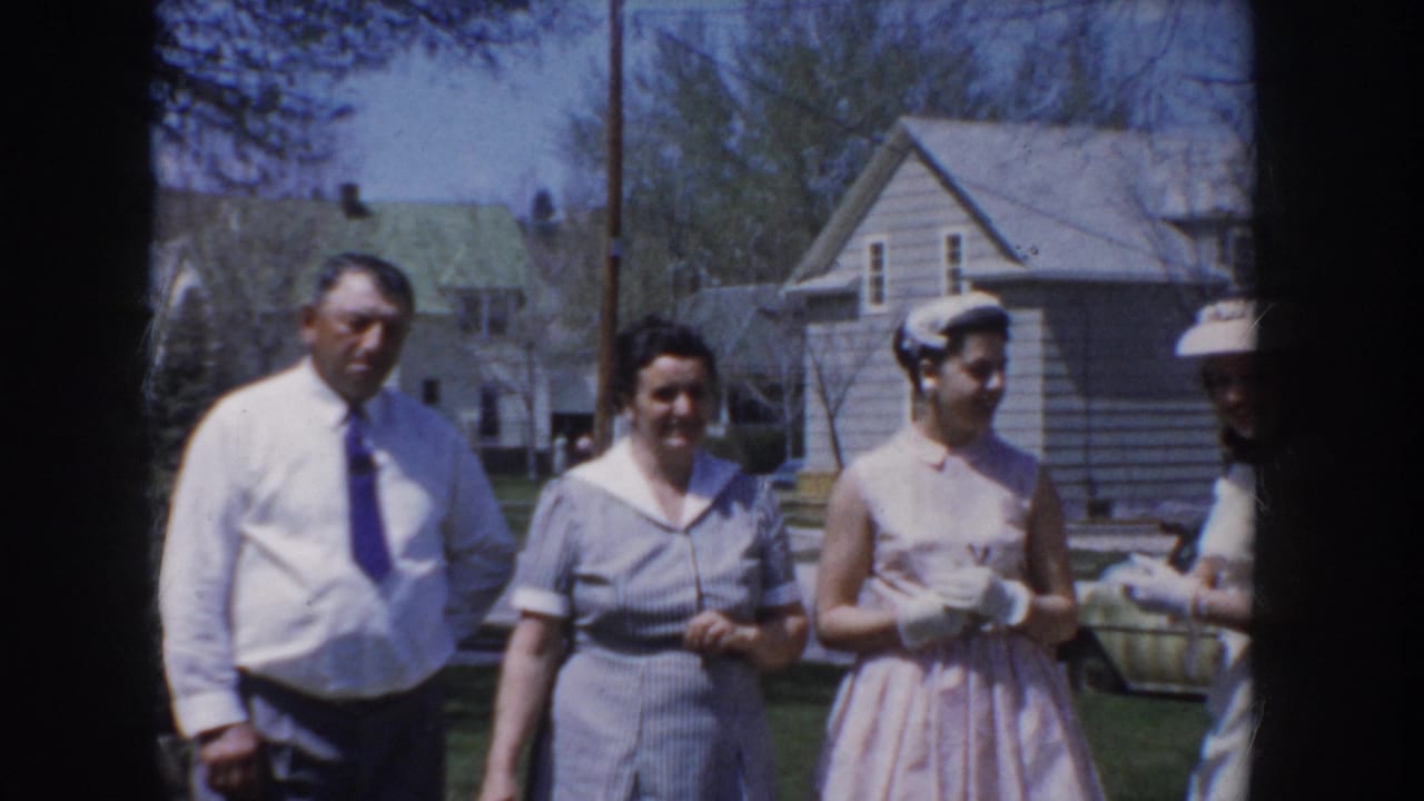 Man in white dress shirt and blue-purple tie poses with three women in pastel mid-1950s dresses and structured hats in a suburban Idaho neighborhood, circa 1955.