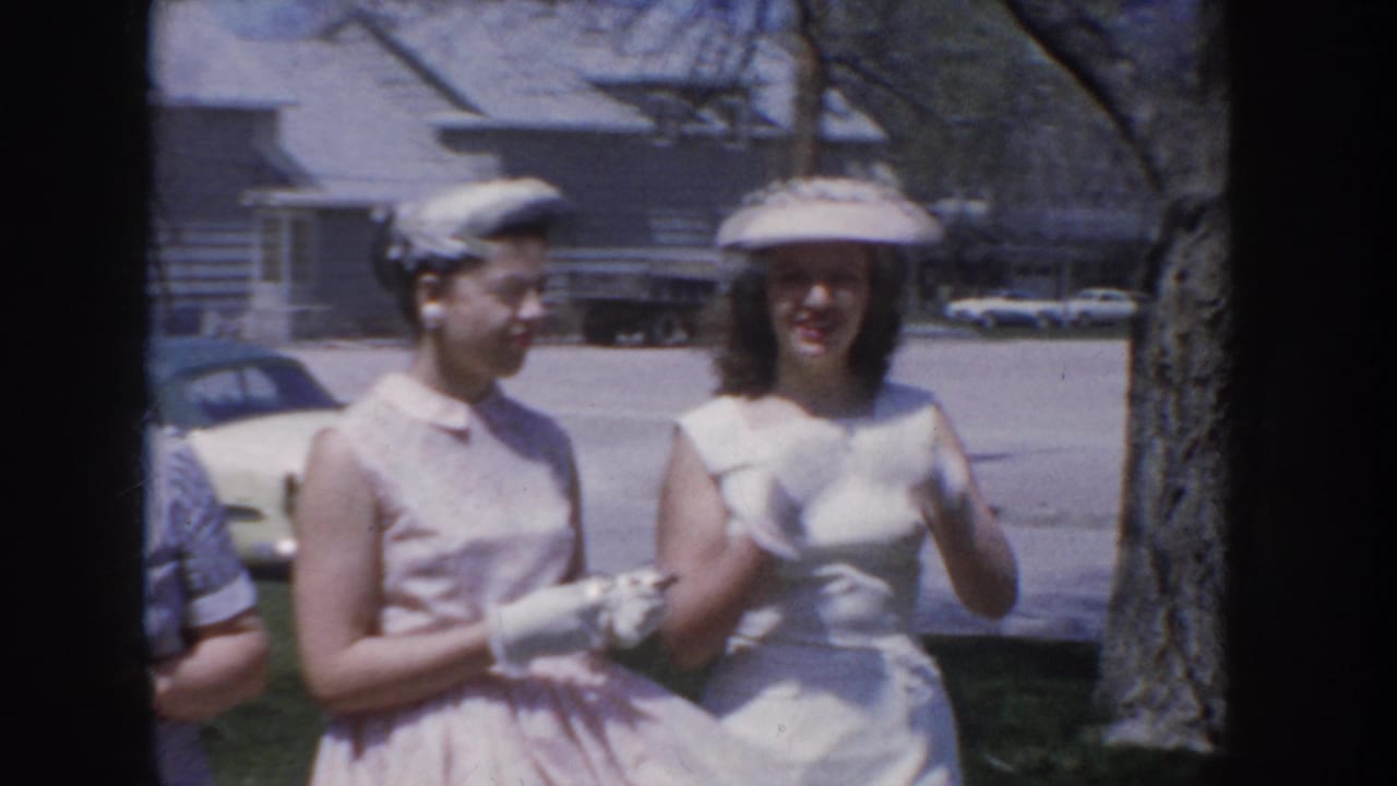 Two women in mid-1950s pastel sleeveless dresses and small structured hats — one holding white gloves — pose outdoors near a parked period automobile in a suburban Idaho neighborhood, circa 1955.