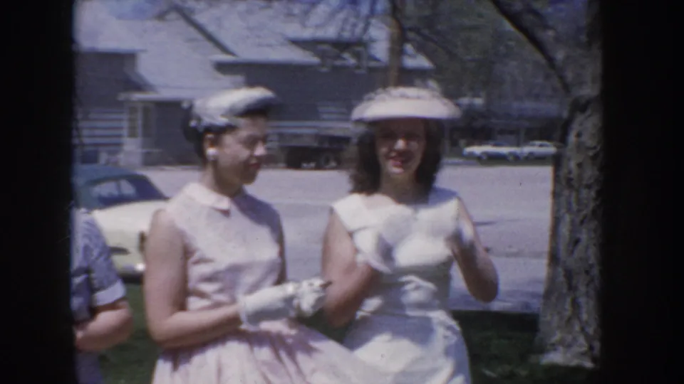 Two women in mid-1950s pastel sleeveless dresses and small structured hats — one holding white gloves — pose outdoors near a parked period automobile in a suburban Idaho neighborhood, circa 1955.