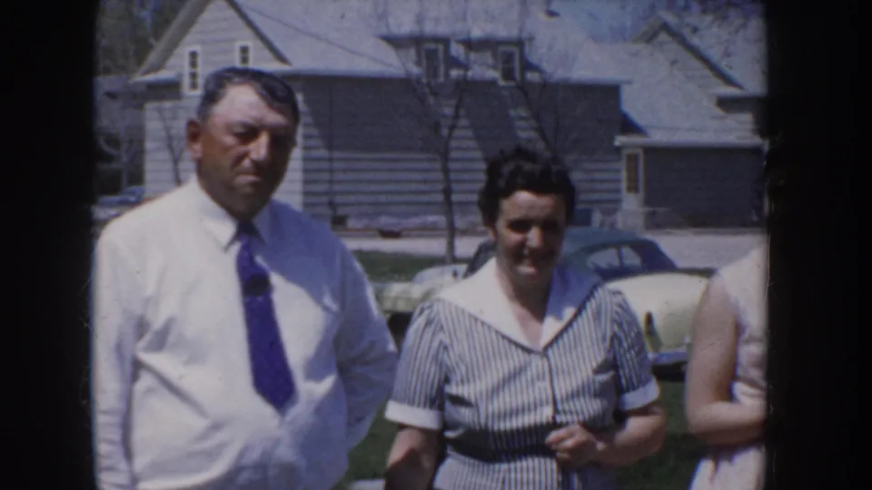 A man in a white dress shirt and blue-purple tie stands with a woman in a grey-striped short-sleeve dress in a suburban residential yard, circa 1955 Idaho.
