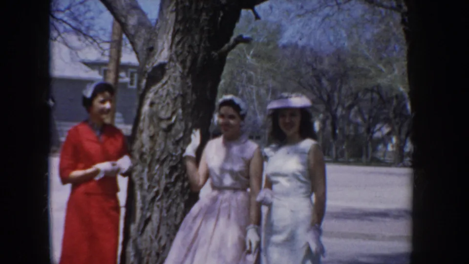 Three women in spring dresses — one in red, two in pastel with small hats and white gloves — stand near a large tree trunk in a suburban Idaho neighborhood, circa 1955.