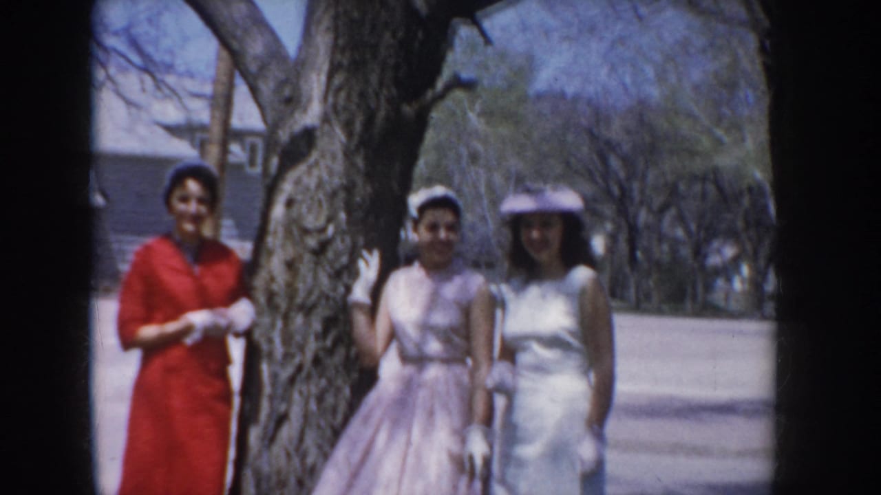 Three women in formal dresses stand near a large bare tree, one in red, two in pastel.