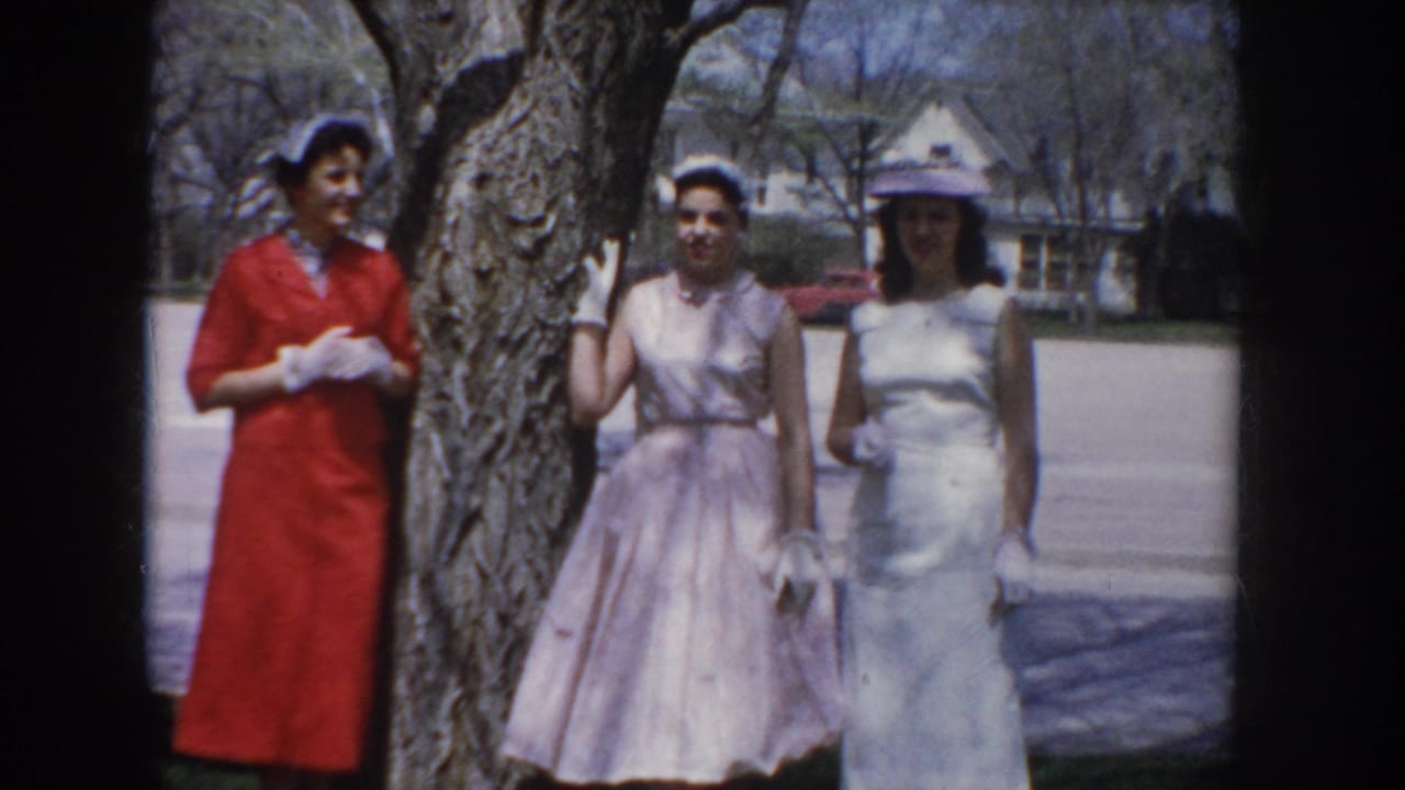 Three women in coordinated mid-century dresses and hats pose together beside a bare tree trunk in a suburban Idaho neighborhood, circa 1955.
