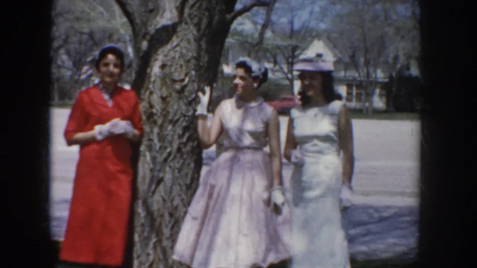 Three women in 1950s formal dresses and hats interact between poses beside a large tree; a red car is partially visible behind them in the suburban street.