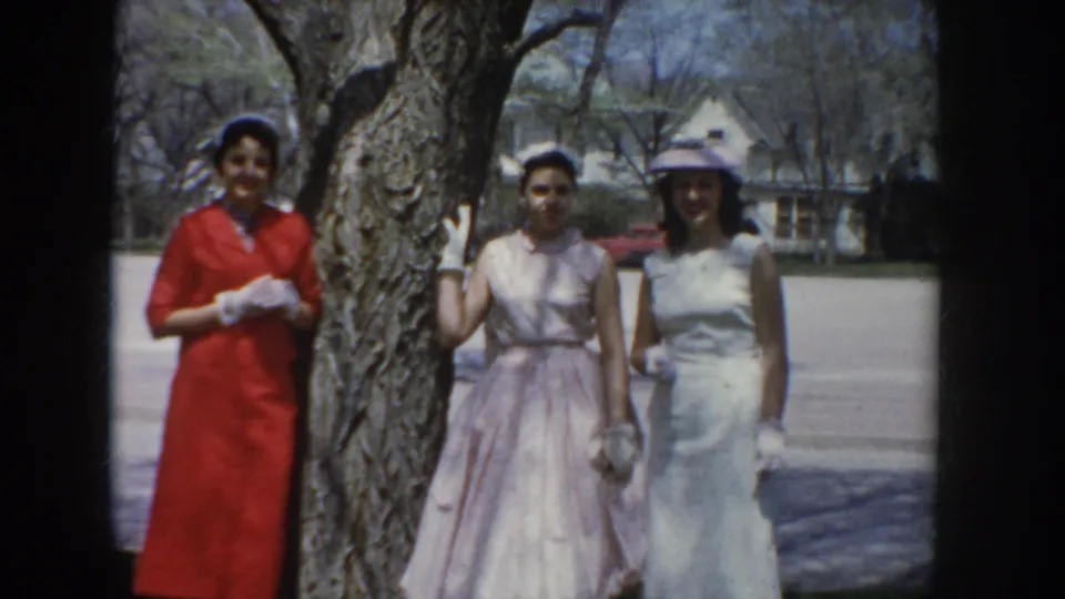 Three women in coordinated mid-century formal attire, including hats and white gloves, pose beside a bare tree with suburban houses and street visible behind them, circa 1955 Idaho.