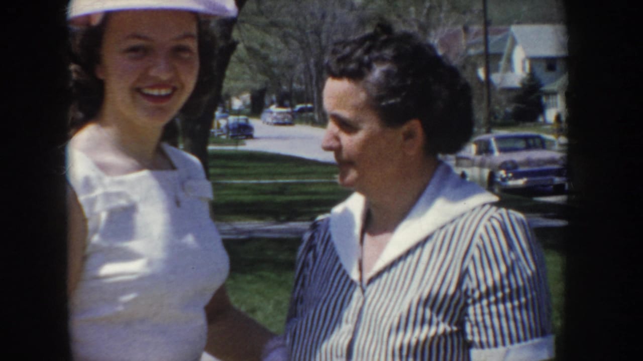Close-up of two women at a 1955 Idaho outdoor gathering; one in a white top and small hat smiles at the camera, the other in a blue striped blouse; period automobiles parked on a suburban street behind them.