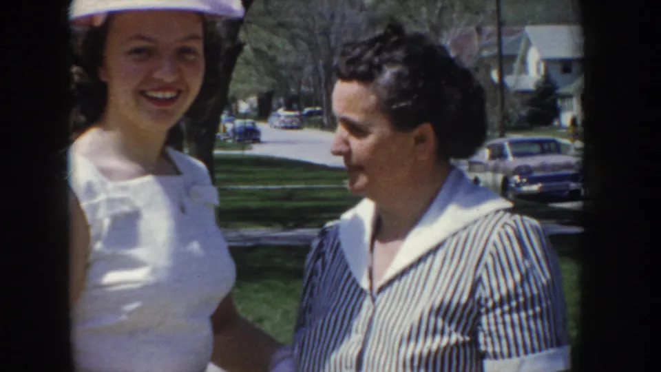 Close-up of two women at a 1955 Idaho outdoor gathering; one in a white top and small hat smiles at the camera, the other in a blue striped blouse; period automobiles parked on a suburban street behind them.