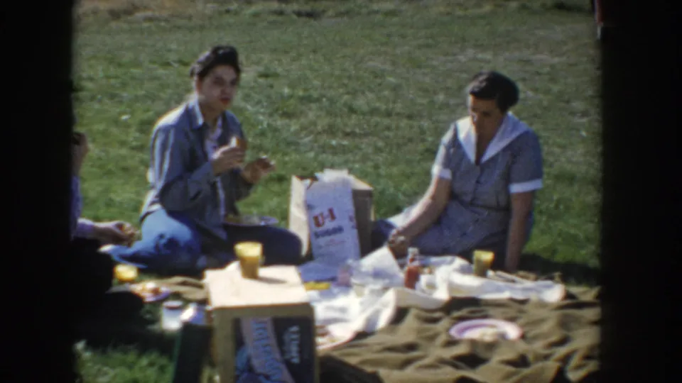 Two women mid-conversation at a 1950s Idaho picnic; a U-I branded paper bag, yellow cups, a condiment bottle, and scattered food fill the foreground of the khaki blanket.