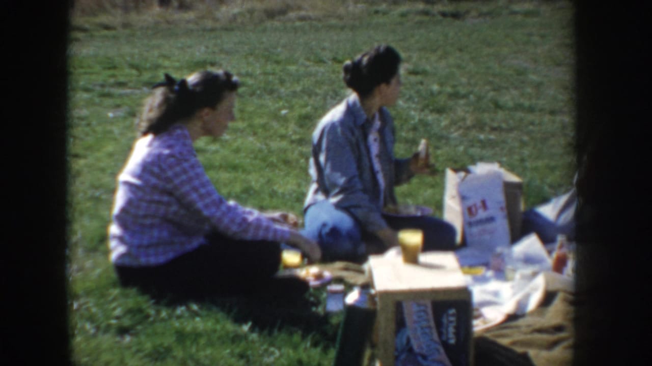 Two women sit on open grassland at a family picnic in Idaho, circa 1955, with a small table holding paper cups and food supplies.