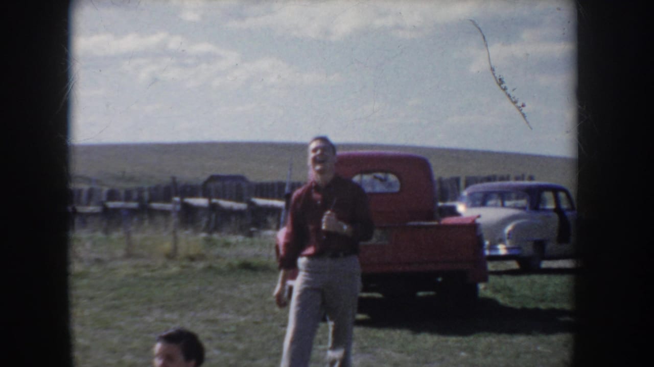 A man laughs toward the camera in an open Idaho field, circa 1955, with a classic red pickup truck and light sedan parked behind him on flat prairie.