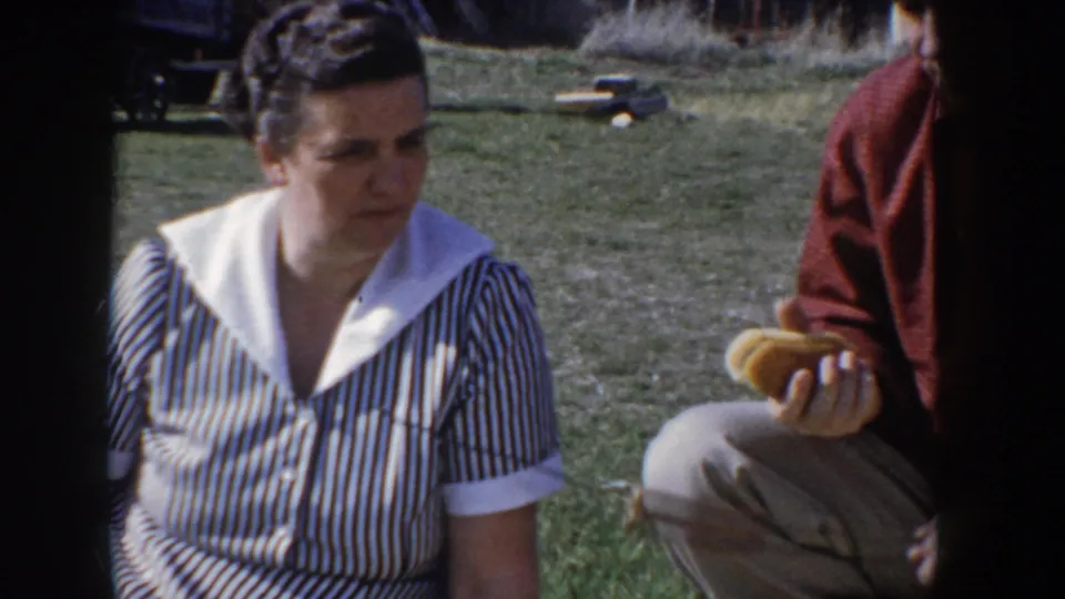 A woman in a striped dress with white collar sits on open grassland at a family picnic in Idaho, circa 1955, with another figure partially visible beside her.