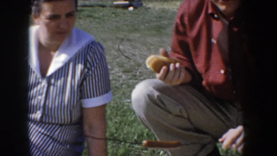 Woman in blue-and-white striped shirtdress and man in red button-down shirt wearing glasses sit cross-legged on grass at a mid-1950s Idaho family picnic; man holds a hot dog bun