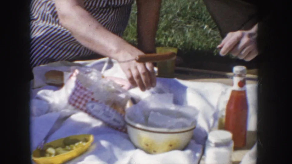 Close-up of a mid-1950s picnic spread on a white blanket: ketchup bottle, yellow serving bowl, white mixing bowl, packaged buns, red-checkered napkin, and condiment jar; woman's hands reach into frame