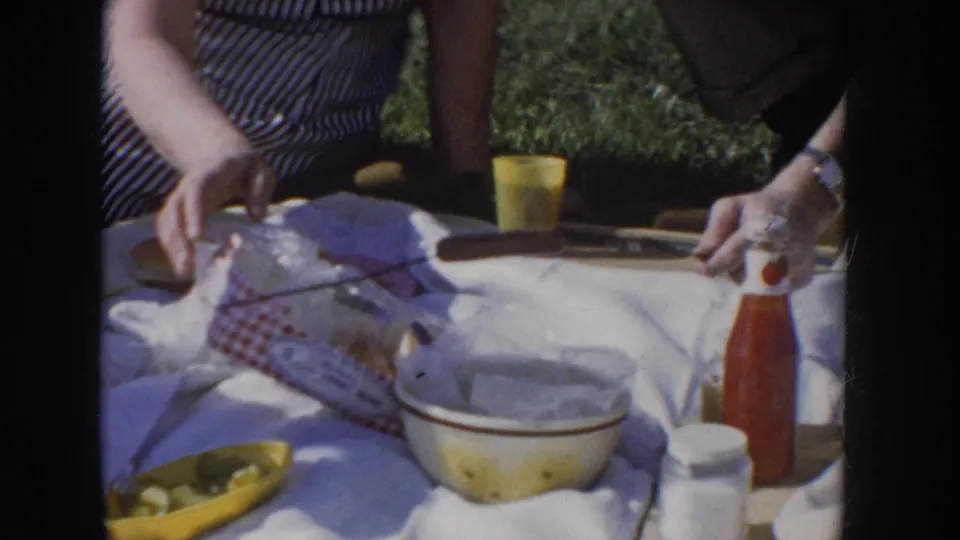 Woman in blue-and-white striped shirtdress leans over a picnic blanket on grass, reaching toward a spread including a yellow tumbler, ketchup bottle, white mixing bowl, and red-checkered cloth