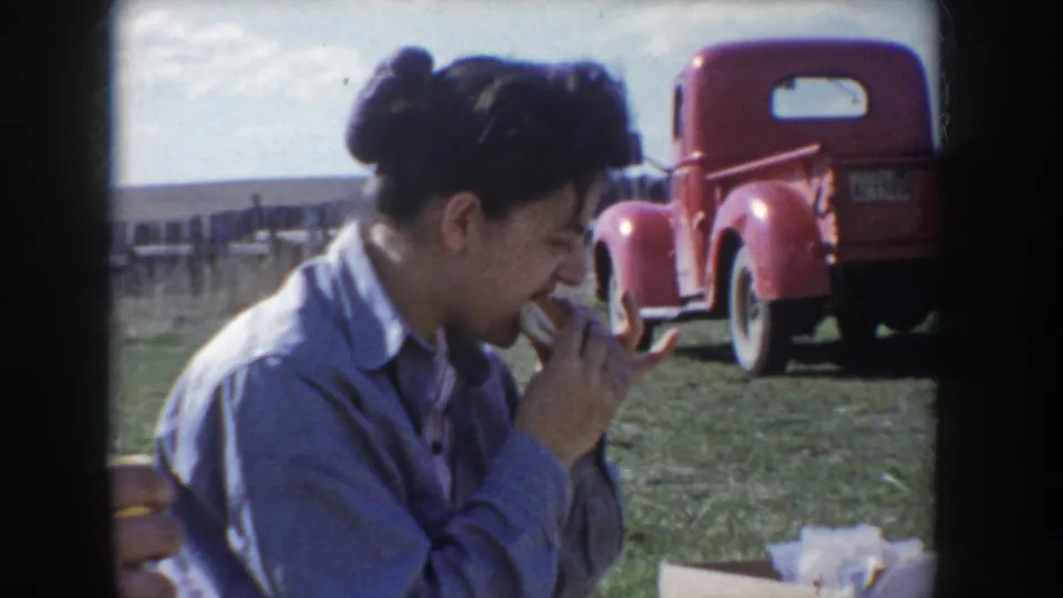 Woman with dark hair in a bun, wearing a light blue shirt, bites into food at a picnic on an open Idaho field; a bright red vintage pickup truck with white-wall tires is parked in the background