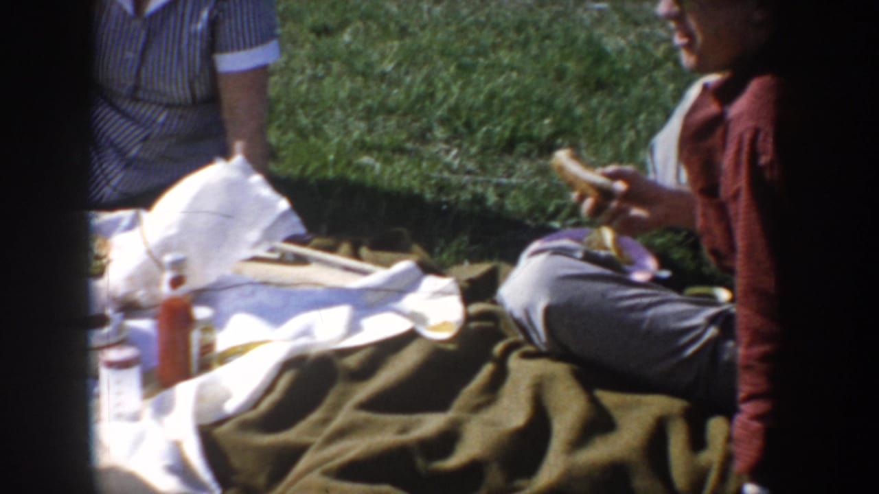 Overhead picnic scene with a man in a red shirt holding food, a white blanket and olive blanket spread on green grass, a red condiment bottle, and the edge of a striped garment at left.