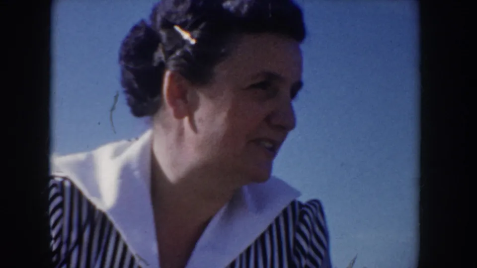 8mm home-movie close-up of a woman in a 1950s striped dress with wide white collar, animated mid-speech expression, at an Idaho outdoor picnic circa 1955, grass and blue sky in background.