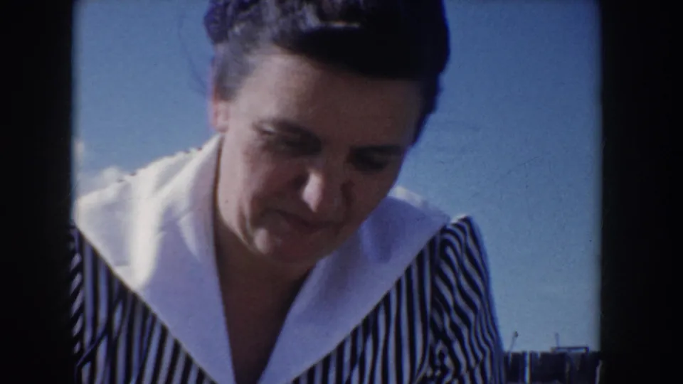 Tight 8mm close-up of a woman in a 1950s black-and-white striped dress with wide white collar, glancing downward with a relaxed expression, at an Idaho outdoor picnic circa 1955, blue sky visible.