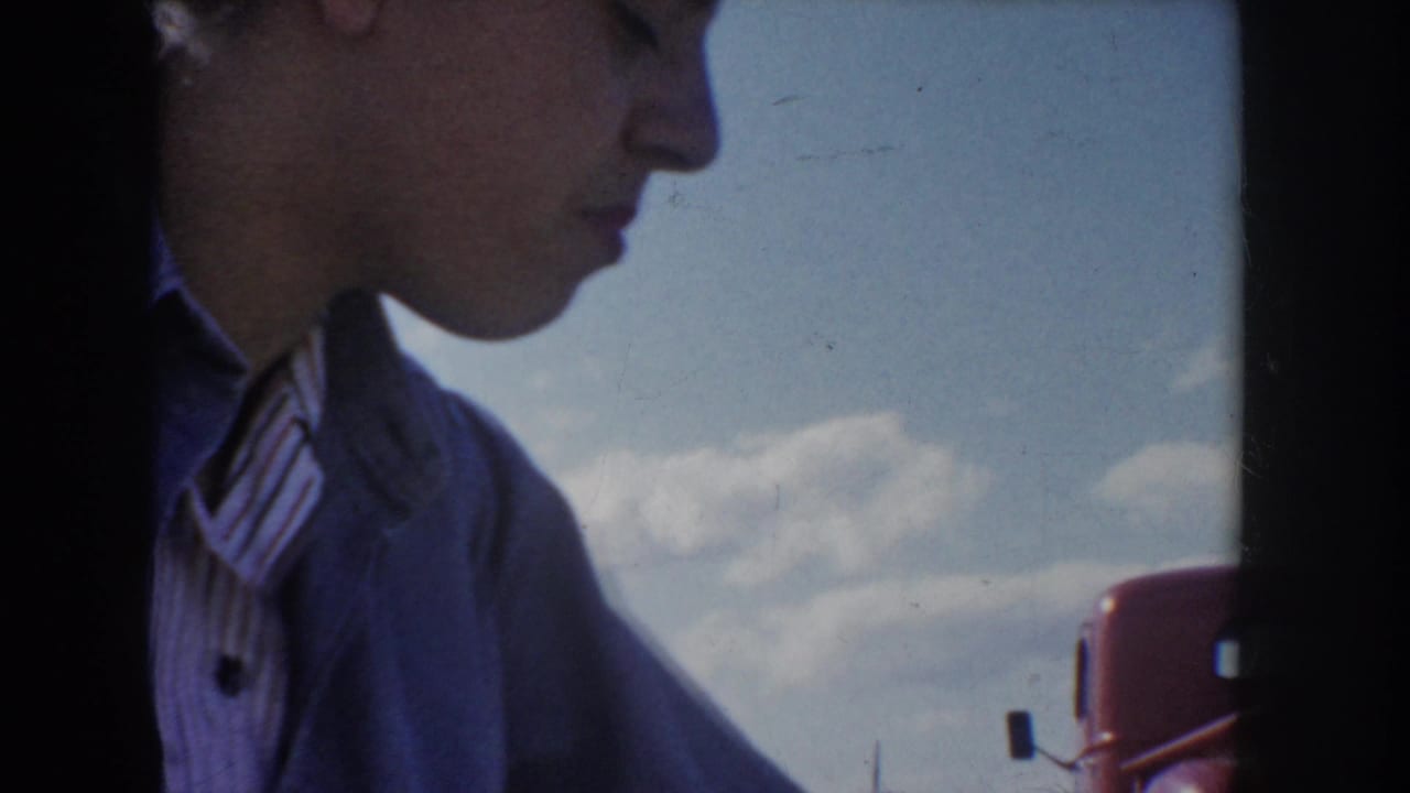 Young woman in light blue jacket and striped collar shirt in close left-facing profile, looking down; blue sky, clouds, and a partial red vehicle visible in background, Idaho USA, circa 1955.