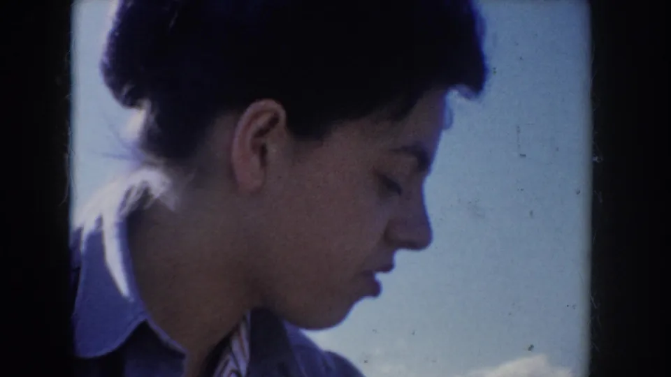 Tight close-up profile of a young woman with short dark hair wearing a light blue jacket, looking downward against a bright blue sky, Idaho USA, circa 1955.