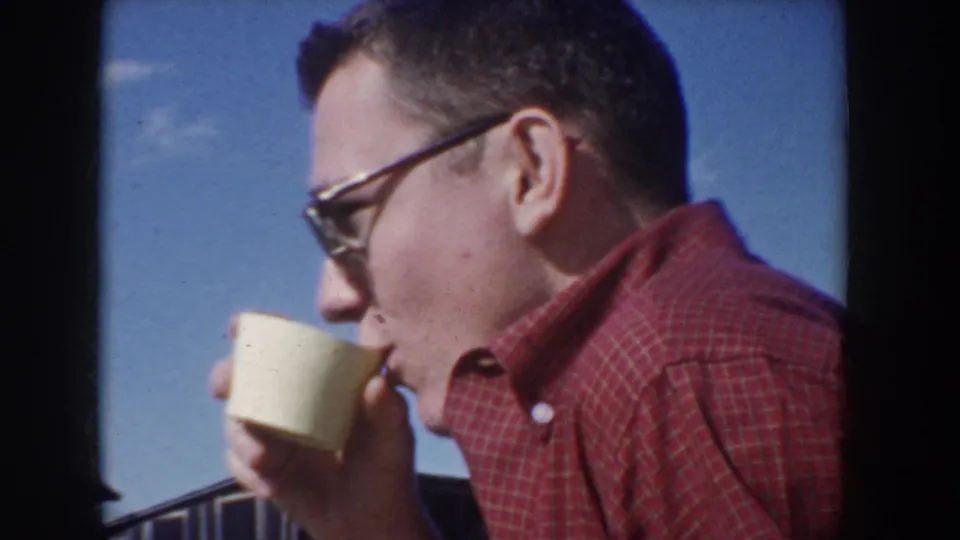 Man in red plaid shirt and 1950s cat-eye sunglasses drinks from a cream paper cup outdoors against a blue sky with scattered clouds, Idaho USA, circa 1955.