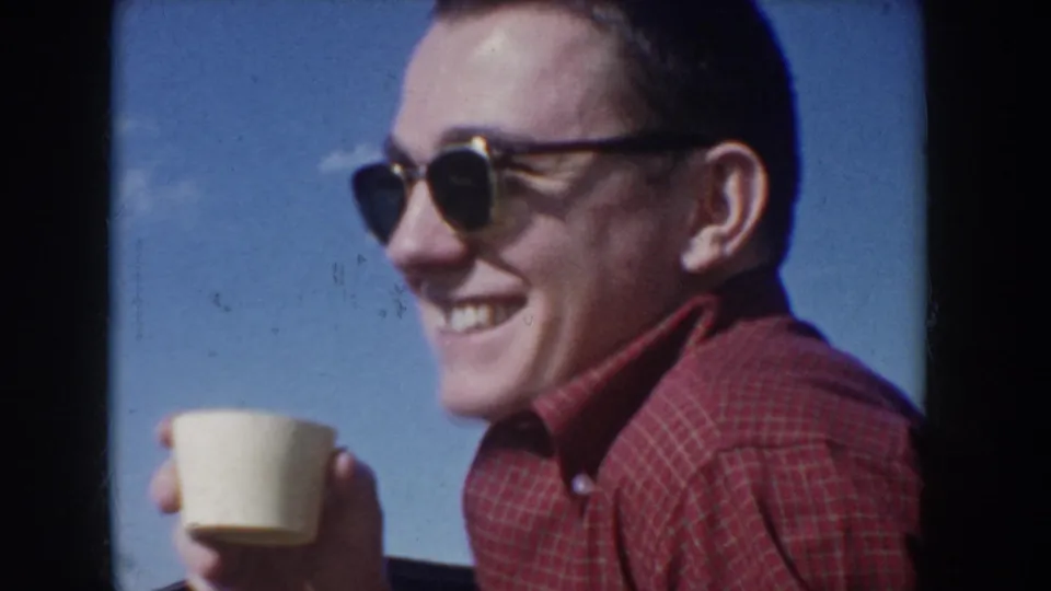 Man in 1950s cat-eye sunglasses and red plaid shirt laughs broadly while holding a cream paper cup outdoors against a clear blue sky, Idaho USA, circa 1955.