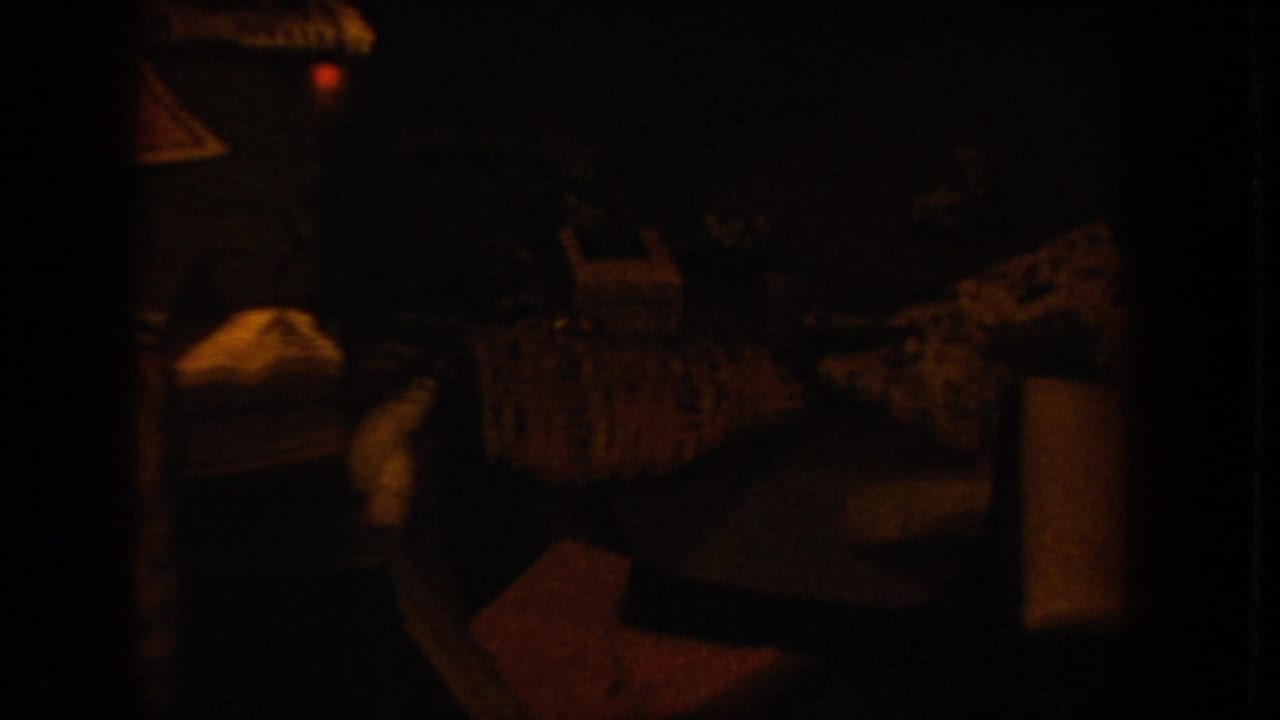 Underexposed 8mm frame showing faint warm-toned shapes; possible stacked luggage or cargo visible amid heavy darkness, with a red indicator light above.